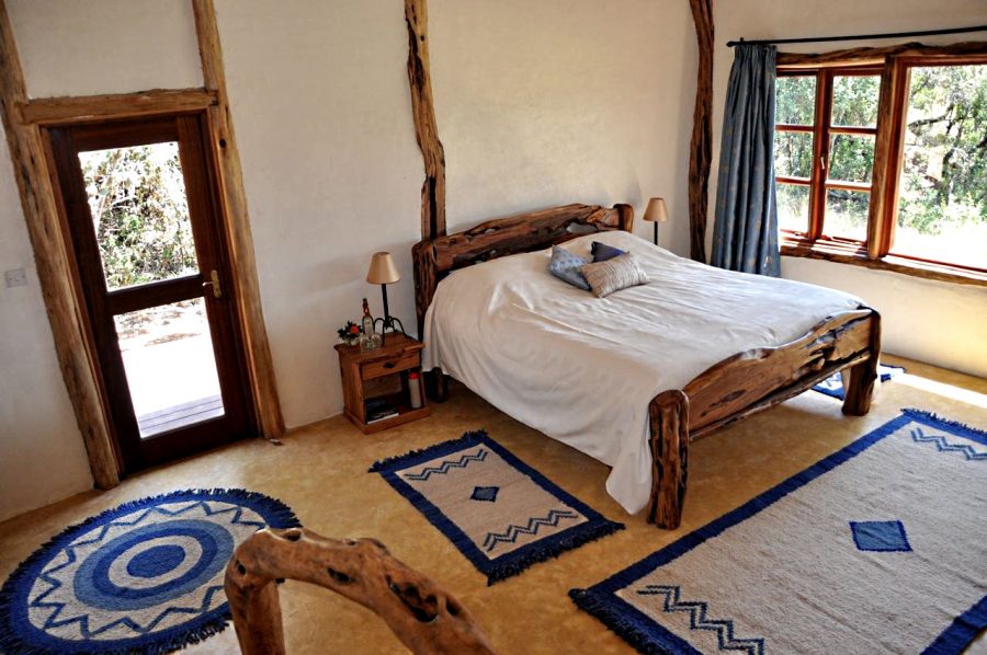 A cozy bedroom with a wooden bed and white bedding, accompanied by decorative pillows. There are two lamps on either side of the bed, a nightstand with a plant, and a window draped with light blue curtains. The floor features circular blue and white rugs, and a wooden door is visible leading outside.