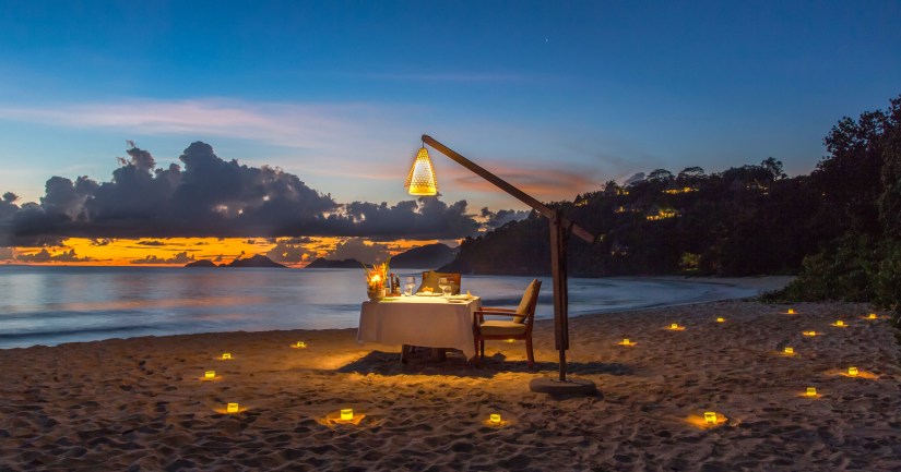A romantic beachside dinner setup at sunset, featuring a table with a white tablecloth, surrounded by glowing candles in the sand, and a lantern hanging overhead illuminating the scene.