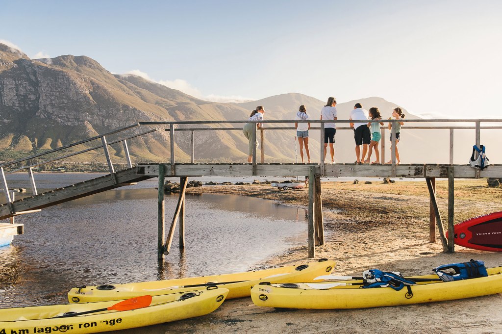 Group of people standing on a wooden pier overlooking a calm lake surrounded by mountains, with yellow kayaks on the sandy shore.