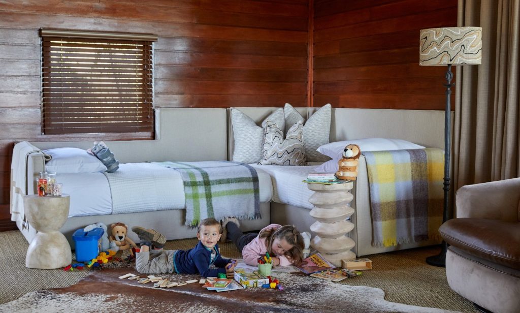 A cozy children's bedroom featuring two beds, with a boy playing on the floor and a girl drawing. The room has wooden walls, a large window with blinds, and is decorated with toys, books, and a standing lamp.