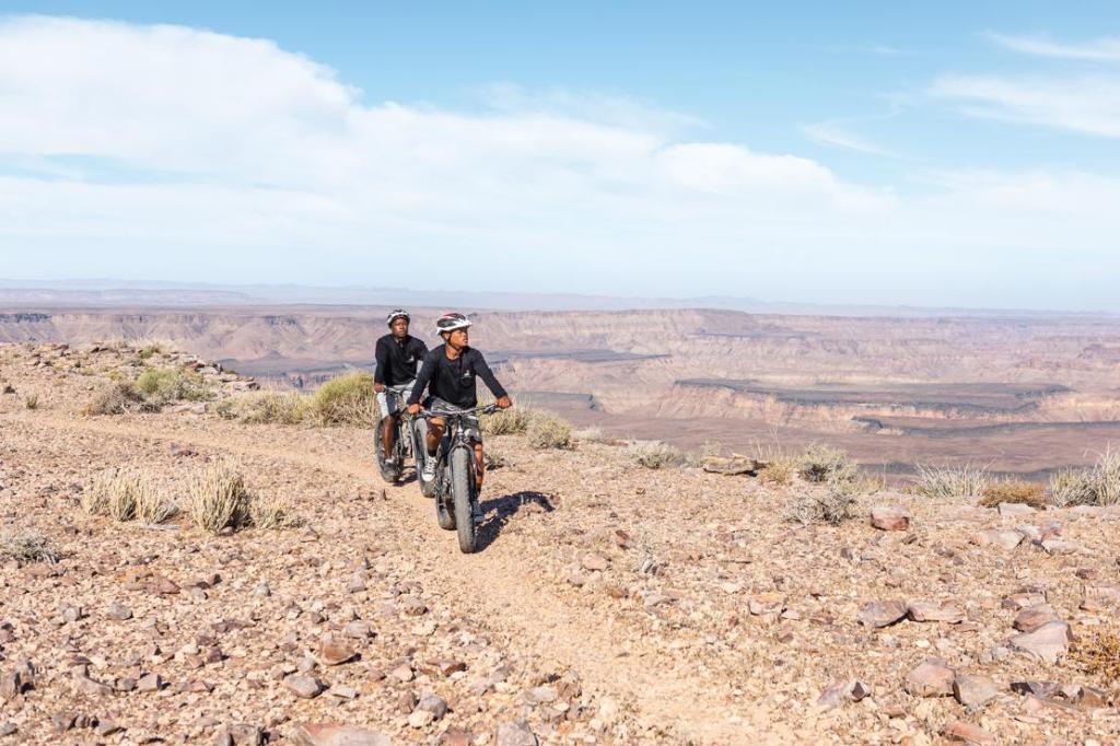 Two mountain bikers riding on a rocky trail in a desert landscape with distant canyons under a blue sky.