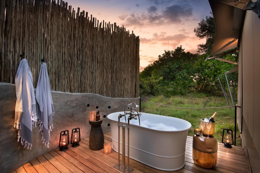 Outdoor bathtub setup with wooden privacy fence, two hanging towels, lanterns, and a scenic view during sunset.