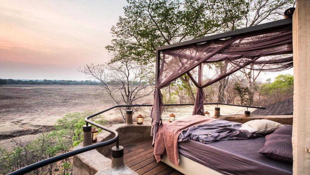 A cozy outdoor bedroom with a bed draped in fabric, surrounded by a natural landscape and trees, during sunset.