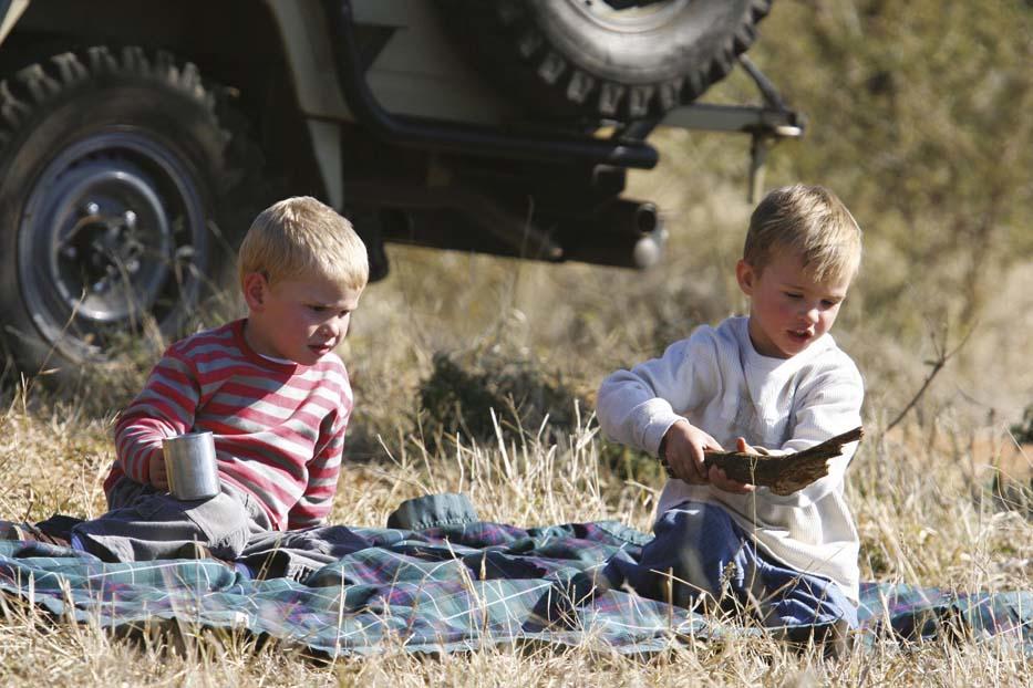 Two young children sitting on a blanket outdoors, one holding a metal cup and the other examining a piece of wood, with a vehicle in the background.